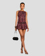A full body view of a brunette model with her legs crossed wearing a sleeveless dress with a maroon and black leaf pattern, featuring a fitted bodice and a flared skirt with a black belt. She is also wearing gold hoop earrings, bangles, black open-toed heels, and holding a cylindrical handbag.