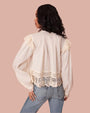 A backview of a model wearing a cream-colored, long-sleeved shirt with intricate lace detailing. The shirt features a pleated back and lace panels along the shoulders and hem. The person is also wearing light blue denim jeans.