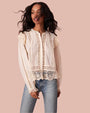 A front view of a model wearing a cream no-show button down blouse with intricate lace detailing and ruffled accents on the shoulders and hem. The blouse is paired with light blue denim jeans.