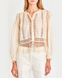 A front view of a model wearing a cream no-show button down blouse with intricate mesh lace detailing and ruffled accents on the shoulders and hem. The blouse is paired with white jeans.