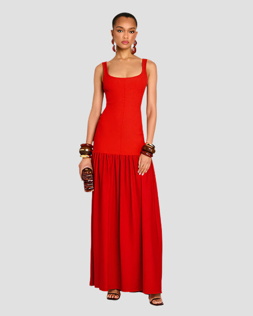 A brunette model standing against a white background, wearing a long, sleeveless red dress with a scoop neck, a fitted bodice and a gathered skirt. The model is holding a small, rectangular clutch in the left hand and is wearing multiple bangles on both wrists, with dangly earrings and brown sandals.