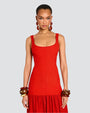 A closer view of a brunette model in front of a white background wearing a long, sleeveless red dress with a ribbed and fitted bodice, and a gathered skirt. The person is wearing multiple bangles on both wrists, with dangly earrings.