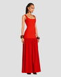 An angled sideview of a brunette model standing against a white background, wearing a long, sleeveless red dress with a scoop neck, a fitted bodice and a gathered skirt. The model is wearing multiple bangles on both wrists, with dangly earrings and brown sandals.
