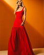 An upwards angle of a blonde model standing against a yellow background, wearing a long, sleeveless red dress with a scoop neck, a fitted bodice and a gathered skirt, paired with gold bangles and a large ring.