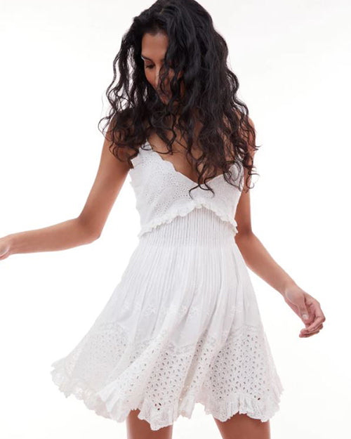 Model wearing a white, sleeveless dress with intricate lace and pleated details, standing against a plain background.