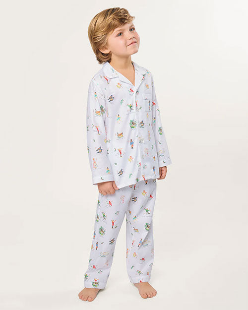 Child standing barefoot against a plain background, wearing a white pajama set with a long-sleeved collared shirt and matching pants, both featuring colorful, whimsical illustrations of animals, plants, and other playful designs.