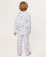 Child standing barefoot with their back to the camera, wearing a light-colored pajama set with long sleeves and pants, decorated with small, colorful illustrations, against a plain background.