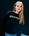 Person with long hair wearing a black sweater with the text 'KISS A COWBOY', standing against a dark background.