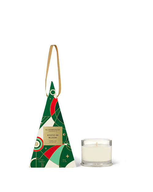 The image features a Glasshouse Fragrances candle next to its packaging. The packaging is designed to resemble a Christmas tree, with a geometric pattern in shades of green, red, and white, and includes the text “Glasshouse Fragrances KYOTO in Bloom Camellia & Lotus.” To the right of the packaging stands a single, lit candle in a clear glass holder. 