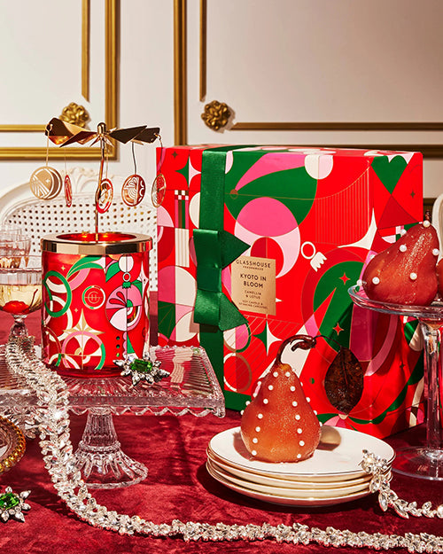 Image showcases a candle and its packaging in a festive holiday table setting featuring a cake stand with a clear stem and base, topped with an assortment of gift boxes in vibrant red and green patterns. The table is covered with a red tablecloth, enhancing the warm, celebratory atmosphere. 