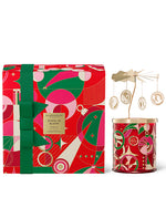 A festive candle with a golden carousel attachment on top is displayed next to its holiday-themed packaging. The packaging features vibrant red, green, and gold geometric shapes and swirls, evoking the holiday season. The candle container is adorned with similar patterns and colors, and the carousel has hanging ornaments that likely spin when the candle is lit, adding a playful touch. The overall presentation suggests it’s designed for a special occasion, possibly as a gift.