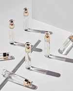 An image featuring seven cylindrical glass perfume bottles arranged haphazardly on a white surface. The bottles have clear liquid inside and are labeled with 'Glasshouse' gold logo and text.