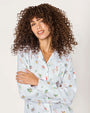  Person with curly hair wearing a white button-up shirt featuring colorful, whimsical illustrations of characters, animals, and plants. They are posing with one arm crossed over the other against a plain background.