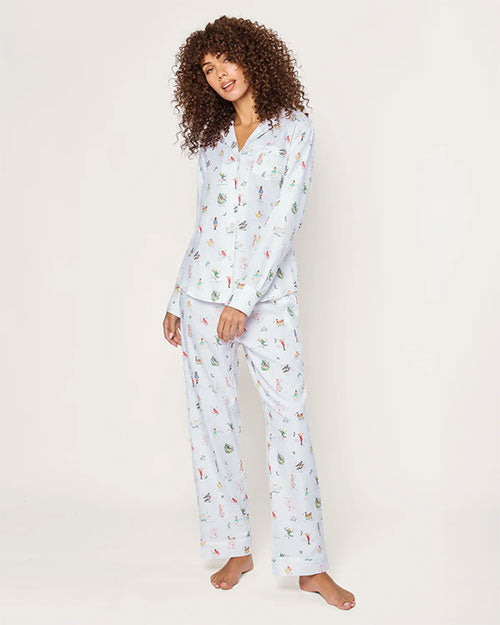Person standing barefoot against a plain background, wearing a light-colored matching pajama set with whimsical, colorful illustrations. The pajama top features long sleeves, a button-down front, and a collar, while the pants are full-length and loose-fitting. The person has curly shoulder-length hair.