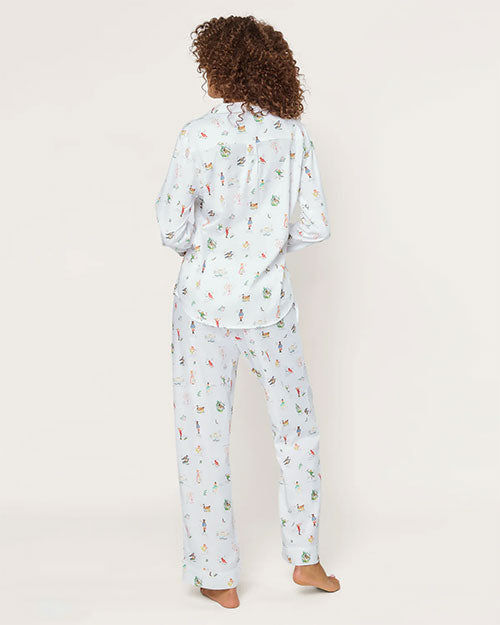 Person standing barefoot with their back to the camera, wearing a white, long-sleeved, button-down pajama top and full-length pants featuring a colorful pattern of small illustrations. The background is plain, and the floor is light-colored.