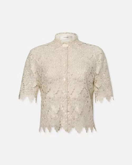 A short-sleeved, lace blouse adorned with a delicate floral pattern. The blouse features a round neckline and a front button closure with small, evenly spaced buttons. Its hem and sleeves exhibit scalloped edges, adding to the intricate detailing. The overall color of the blouse is a neutral, light hue, suggesting an elegant and feminine style. 