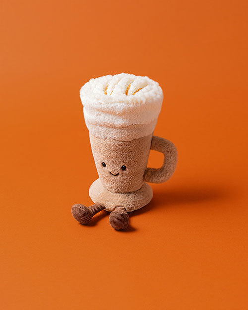 Plush toy shaped like a latte with foam on top, featuring a handle, smiling face with black eyes, and small legs with brown shoes, set against an orange background.