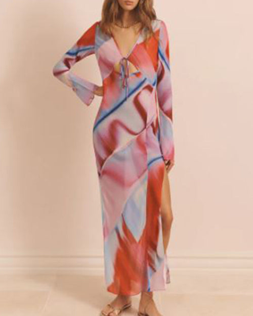 A brunette model with her hand on her hip, wearing a long-sleeve mesh dress with a vibrant abstract multicolored print, a V-neck, an open design under the chest with a bow at the center of the chest, and a side leg slid. The background is plain white.