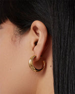 Close-up of an ear wearing a shiny gold hoop earring with a smooth, curved design; straight dark hair partially covers the ear.