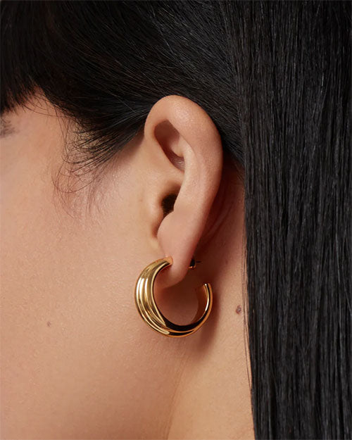 Close-up of an ear wearing a shiny gold hoop earring with a smooth, curved design; straight dark hair partially covers the ear.