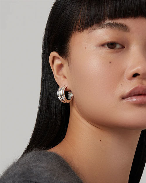 Close-up of an ear wearing a silver hoop earring; person has long, straight black hair, and is wearing a grey top against a plain background.