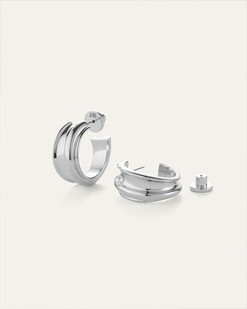 Pair of silver hoop earrings with a modern, chunky design; one earring stands upright showing the post and 'JB' engraved on the back, while the other lies down with its back detached beside it.