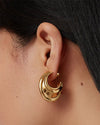 Close-up of an ear wearing a gold hoop earring with a crescent-like design featuring two overlapping curves; dark hair visible at the top edge.