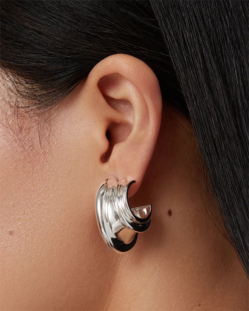 Close-up of an ear wearing a shiny silver hoop earring with a unique, layered wave-like design.