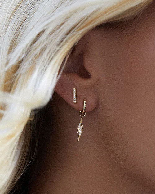 Close-up of a person’s ear adorned with two gold earrings. The upper earring is a small bar with pave diamond and the lower earring is a hoop with a dangling lightning bolt charm. 