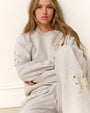 Person sitting against a white wall wearing a light gray sweatshirt and sweatpants adorned with lace bows appliqués on the sleeves and legs.