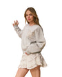 Person standing against a white background, wearing a light gray  sweatshirt and a white layered lace skirt.