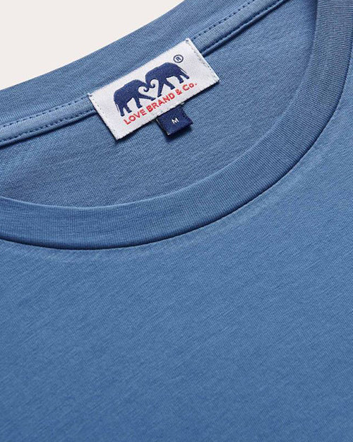 Close-up of a blue crew neck t-shirt with a small white and navy rectangular label stitched at the center of the collar. The label features an elephant silhouette and logo lettering.