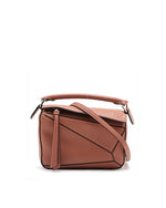 A Loewe brand bag in a warm brown leather, featuring a structured, rectangular shape with rounded corners. It’s viewed from an angle that highlights its depth and dimension, with detailed stitching along the edges, an embossed Loewe logo on the front, and an adjustable shoulder strap with silver-tone hardware.