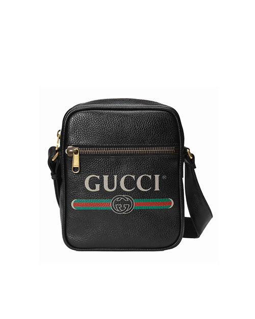 A black Gucci crossbody bag with a textured leather exterior. The front of the bag features a gold-toned zipper pocket with the iconic green and red stripe below it. Above the stripe, the Gucci logo is prominently displayed in gold lettering with the double G emblem centered beneath it. The bag has an adjustable black strap attached to it with gold-toned hardware, suitable for crossbody wear.
