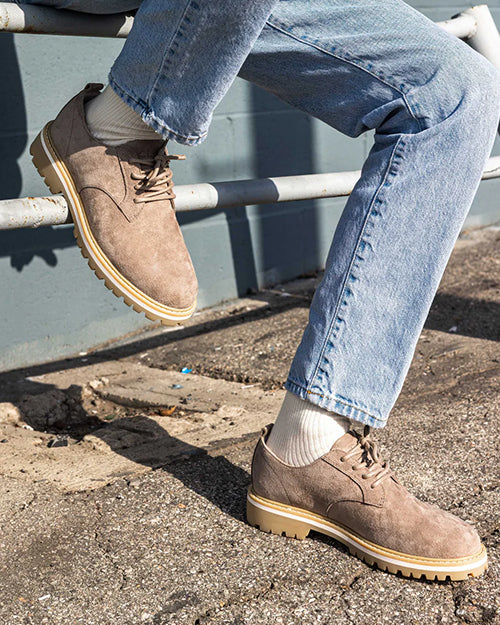 Person wearing brown suede shoes and blue jeans on a concrete surface.