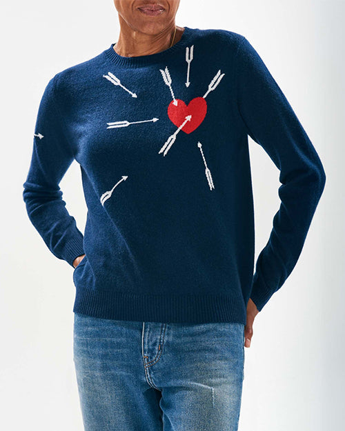 A model wearing a navy-blue sweater with a design of white arrows and a red heart. The arrows are pointing towards the heart, which is located on the left side of the chest area. The sweater is paired with blue jeans.