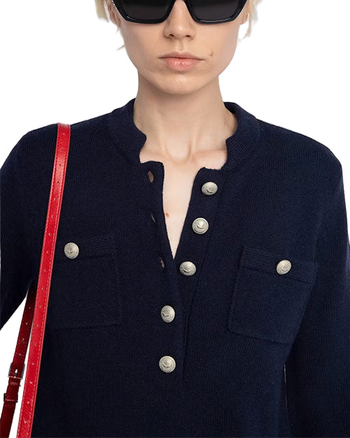Person wearing dark sunglasses, a dark-colored sweater with metallic buttons down the front and two chest pockets with buttons, carrying a red strap over the left shoulder.