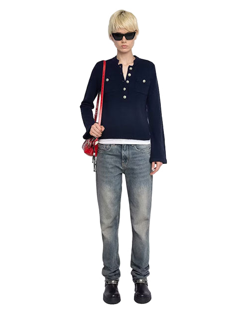 Person standing and facing forward, wearing dark sunglasses, a dark long-sleeve shirt with front buttons, light blue jeans, black shoes, and carrying a red shoulder bag. 