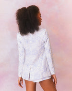 A back view of a model in a white blazer with a subtle blue paisley pattern. The blazer has a tailored fit with a notched lapel and a single-button closure, paired with matching shorts and top. 