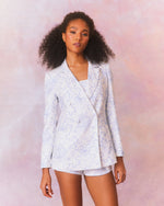 A model in a white blazer with a subtle blue paisley pattern. The blazer has a tailored fit with a notched lapel and a single-button closure, paired with matching shorts and top. 