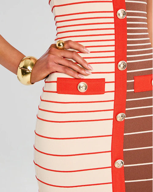 A close-up waist level view of a model wearing a dress with a fitted silhouette. The dress is half white/cream and half brown and features horizontal stripes in varying shades of red and white, with a vertical red stripe running down the center front. The red stripe is adorned with medium sized buttons.  Additionally, the person has gold bracelets on both wrists.