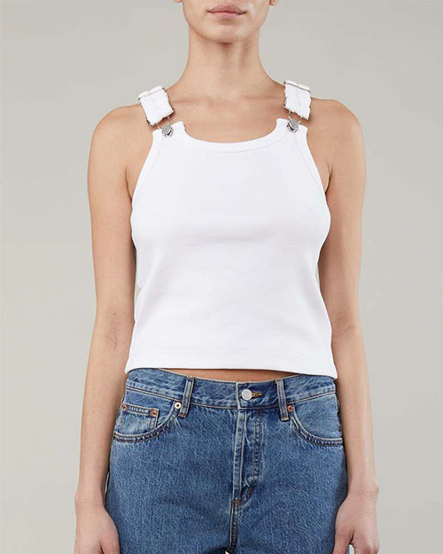 A torso only shot of a model wearing a slightly cropped white tank top with adjustable buckle straps and blue jeans. The background is grey.