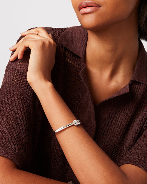 A close-up of a sleek silver bracelet on a person’s wrist. The minimalist design features a thin band with a small knot detail in the center.