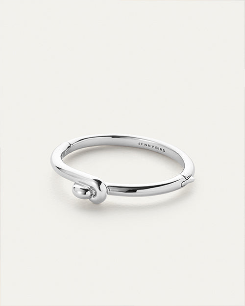 A sleek, silver bracelet with a minimalist design, featuring a smooth band and a small knot detail near the opening. The brand name ‘JENNY BIRD’ is engraved on the outer surface of the band.