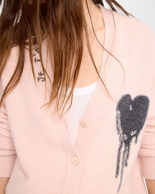 Close-up of a light pink buttoned cardigan worn over a white top. The cardigan features a black heart graphic with a dripping effect on the left side and stylized black text near the neckline on the right side. The buttons are light beige, and the fabric appears soft and knitted.