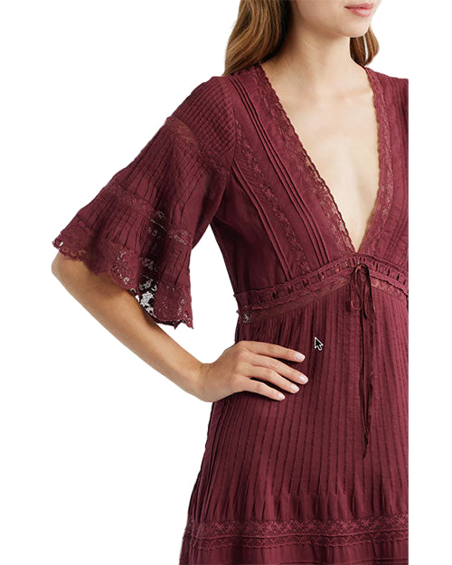 Person wearing a burgundy dress with a deep V-neckline, flared sleeves featuring lace inserts, and pleated fabric. The face is partially obscured, with one hand resting on the hip.
