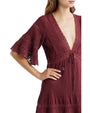 Person wearing a burgundy dress with a deep V-neckline, flared sleeves featuring lace inserts, and pleated fabric. The face is partially obscured, with one hand resting on the hip.