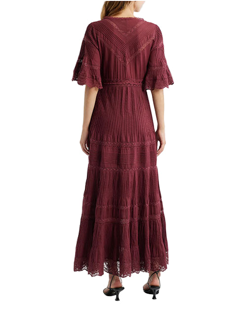 Back view of a person wearing a long burgundy dress with short lace-trimmed sleeves, pleated and lace details, a V-shaped pattern on the upper back, and lace at the hem, paired with black heeled sandals.