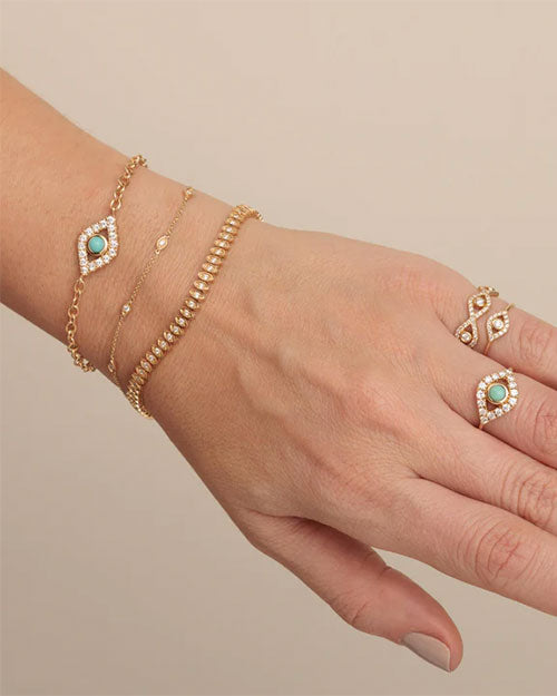 Hand wearing three gold-toned bracelets—one with a turquoise stone and diamonds, one with evenly spaced gold beads, and one with tiny diamond accents—along with two rings, one matching the turquoise bracelet and the other featuring an intricate diamond design.
