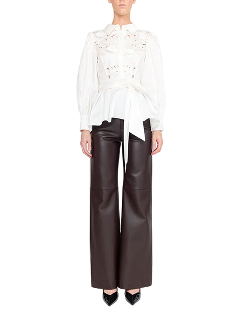 A person is wearing a white long-sleeve blouse with intricate lace detailing on the upper chest and shoulders, paired with high-waisted, wide-leg black leather pants. The blouse has a tied waist and the person is also wearing black pointed-toe shoes.